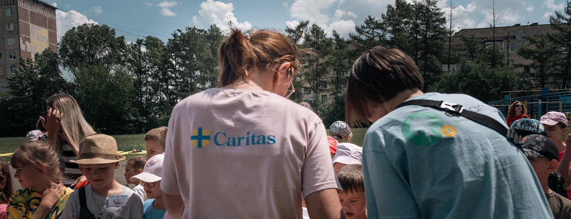 Ukraine-Krieg: Kolleginnen einer Caritas-Partnerorganisation bei einem Sommercamp für Kinder. Zwei Frauen mit Caritas-T-Shirts sind von hinten zu sehen, vor ihnen ein Tisch mit Lebensmitteln. Im Hintergrund sind Kinder zu sehen, die sich etwas zu essen holen.