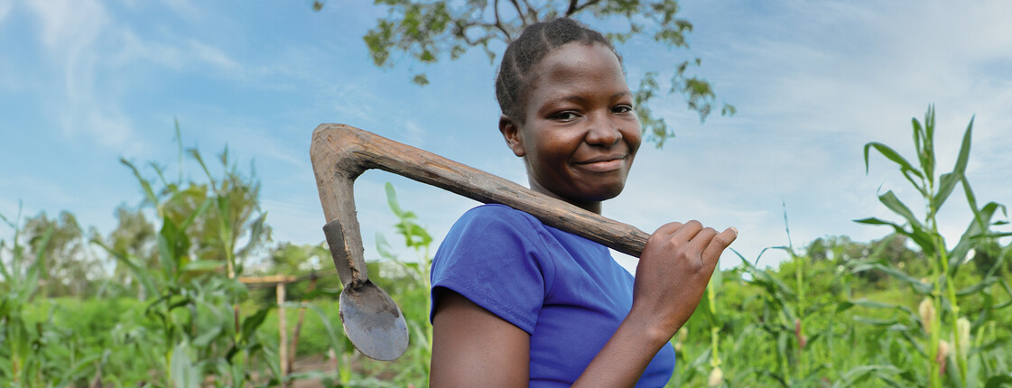 © Nyokabi Kahura_fairpicture Eine junge Frau steht auf einem grünen Feld und trägt eine Hacke über ihrer Schulter. Sie lächelt in die Kamera. Das Bild steht im Kontext der Hungerkampagne 2024.
