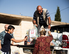 Ein Mann steht auf der Ladefläche eines kleinen LKWs und gibt Säcke mit Gewand an Menschen unten weiter. Auf den Säcken ist das Logo von Caritas Syrien.