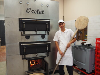 Ein junger Mann mit Brotschieber rechts vor einem großen Backofen stehend