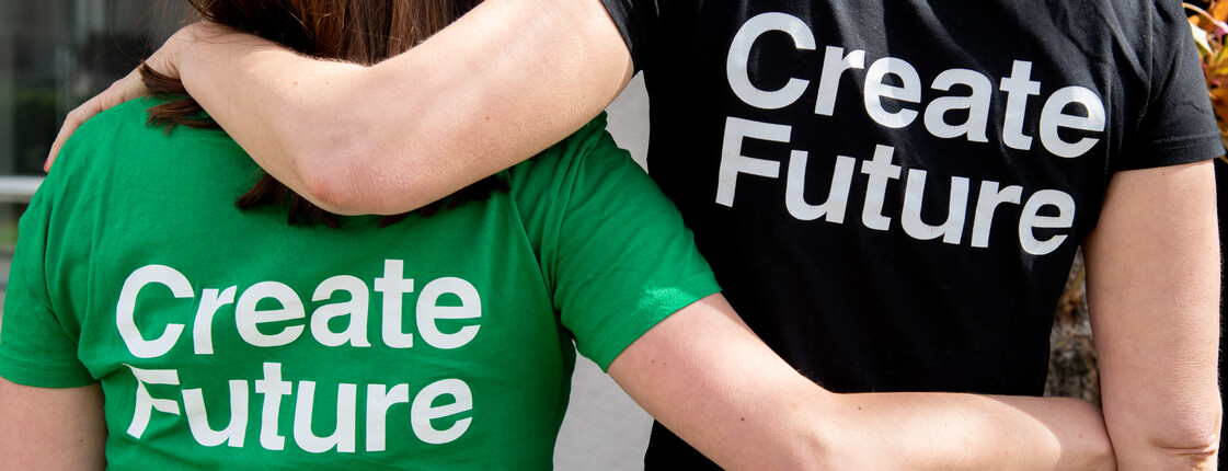Create Future Man sieht zwei Personen von hinten, sie haben die Arme auf ihren Schultern und tragen T-Shirts mit der Aufschrift "Create Future".